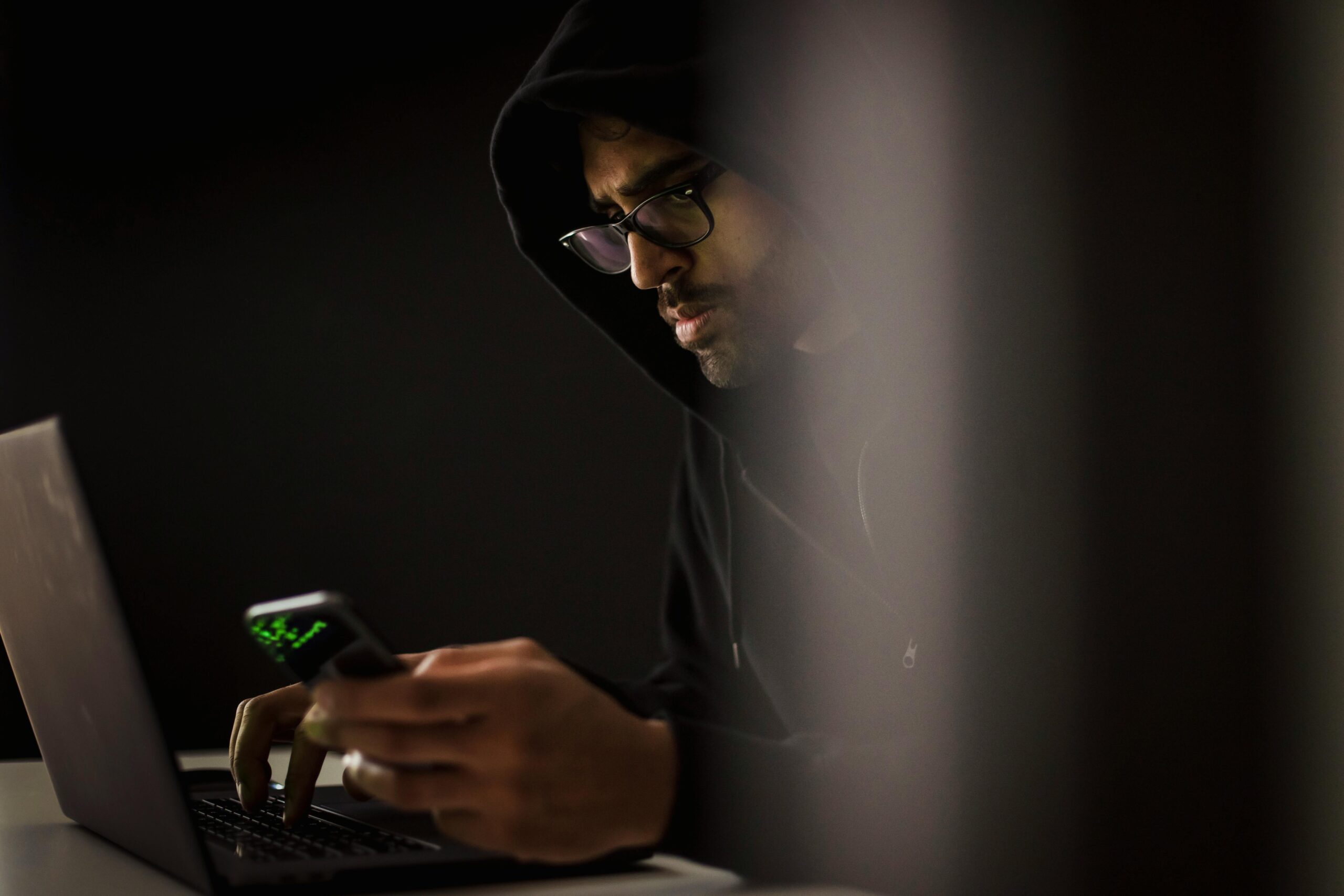 Man in hoodie using laptop and smartphone in dark room, focused and concentrated.