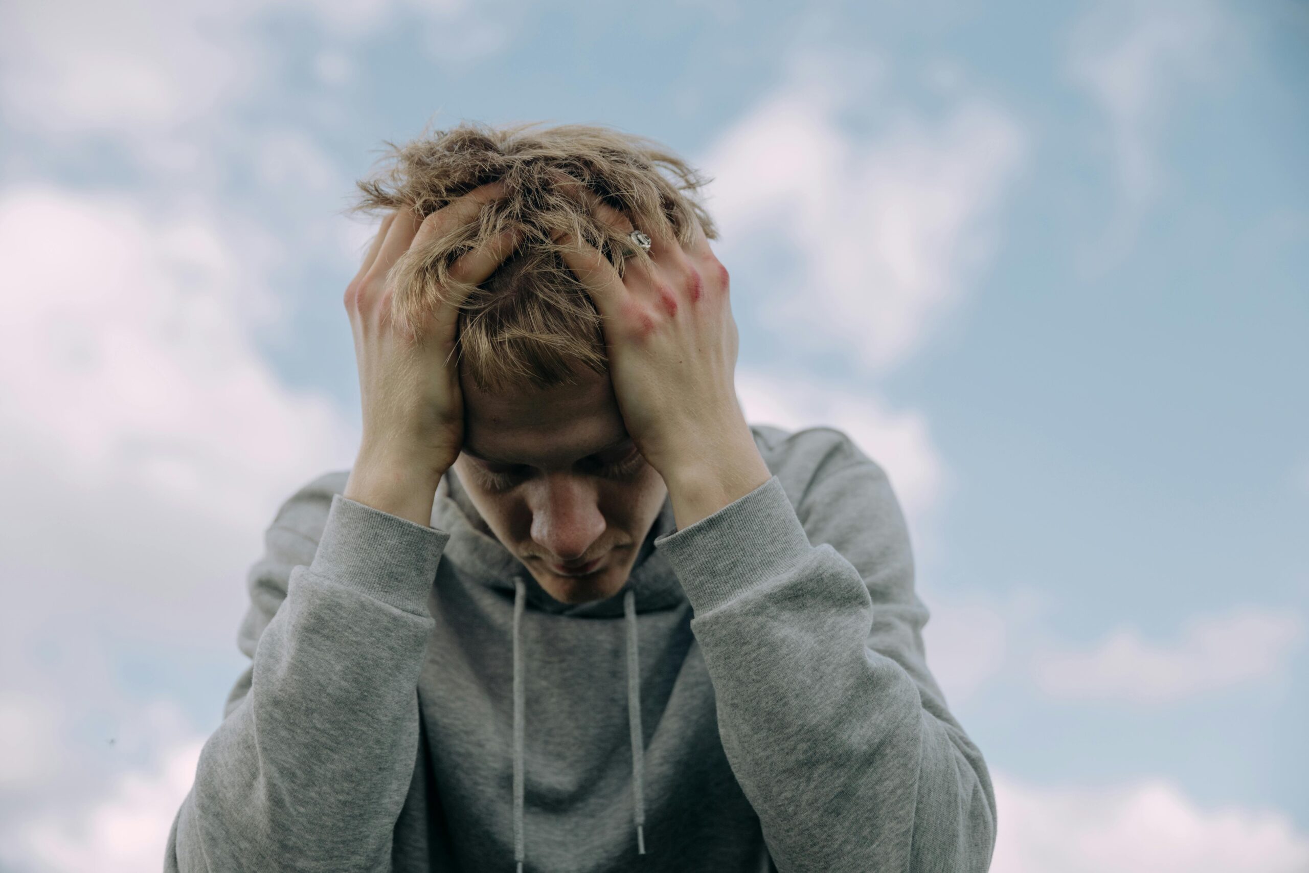 Young man in gray hoodie holds head in frustration, set against cloudy sky.