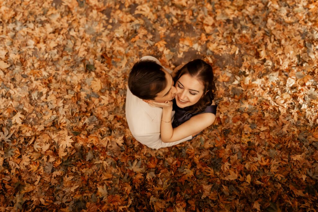 A loving couple embraces amidst fallen autumn leaves, viewed from above.