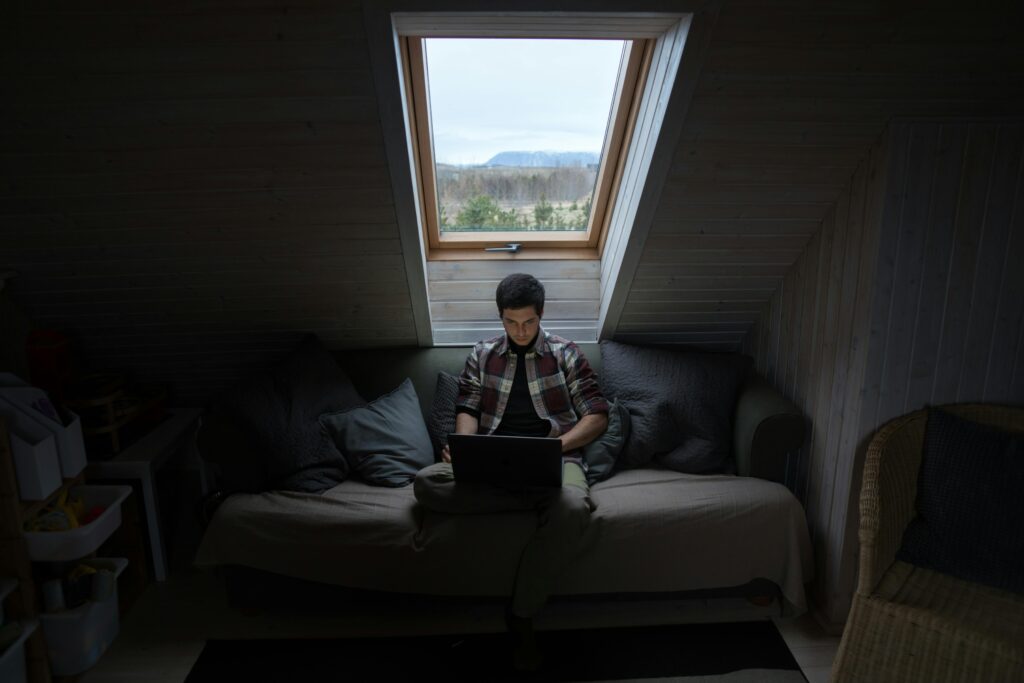 A young man focuses on his laptop in a dimly lit attic room with a window view.