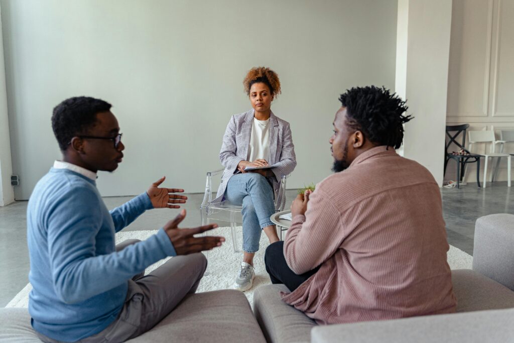 A therapist mediates a session with two men on a couch discussing mental health.